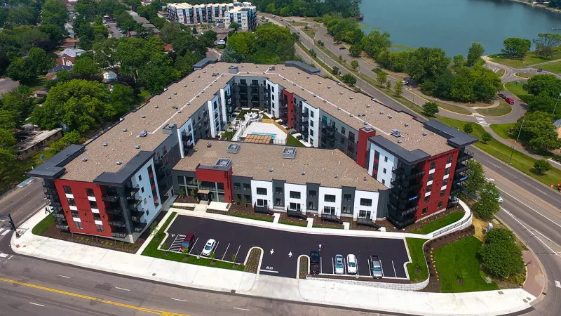 Angled aerial view of a modern, U-shaped apartment complex with red, dark gray, and white sections, a parking lot, and a lake.