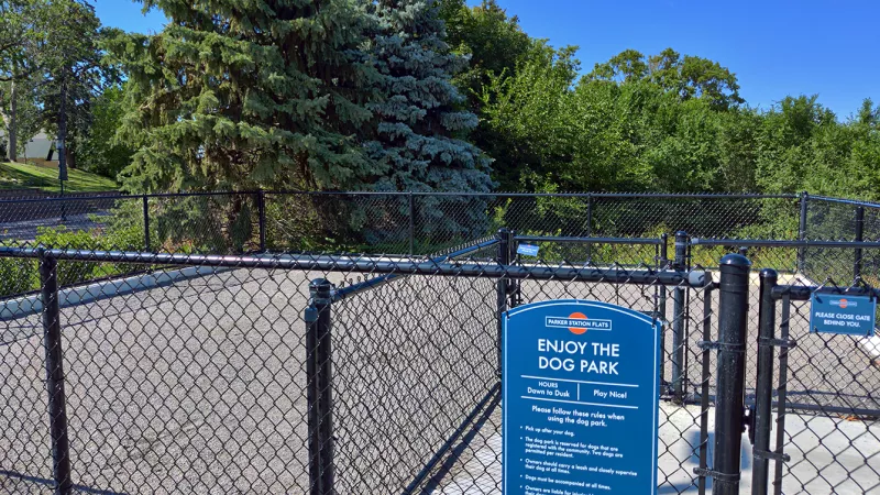 Photograph of a dog park enclosure with a chain-link fence, a blue rules sign, and green trees under a clear sky.