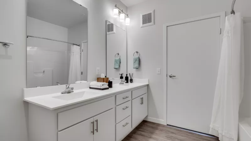 Bright modern bathroom with white double vanity, large mirror, wood-look floor, and white shower curtain.