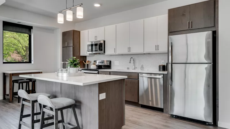 Modern kitchen with a white island, two bar stools, light and dark wood cabinets, and stainless steel appliances.