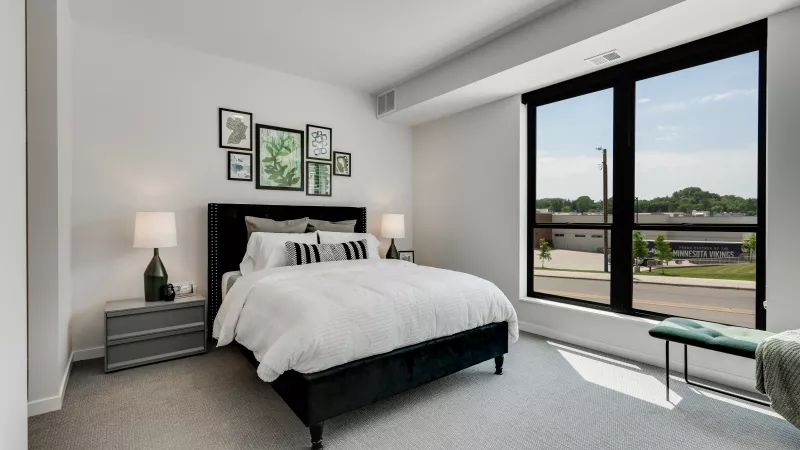 Bright modern bedroom with a dark bed, white duvet, green botanical art, and a large window overlooking a street.