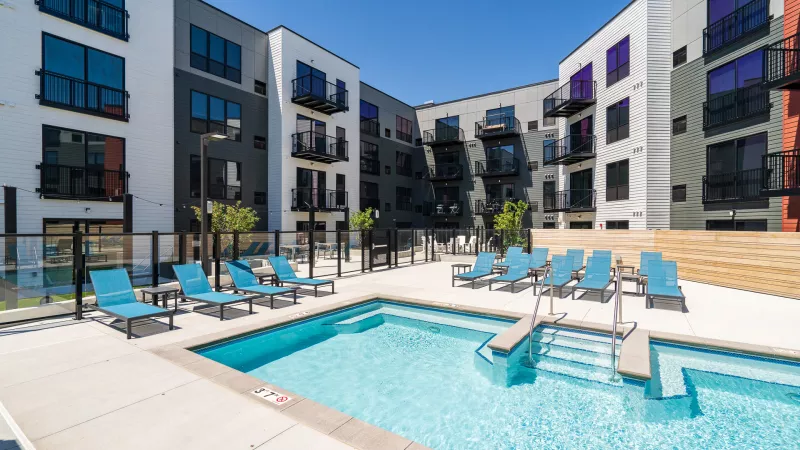 Sunny outdoor pool with turquoise lounge chairs and a hot tub, surrounded by a modern, multi-story apartment building.