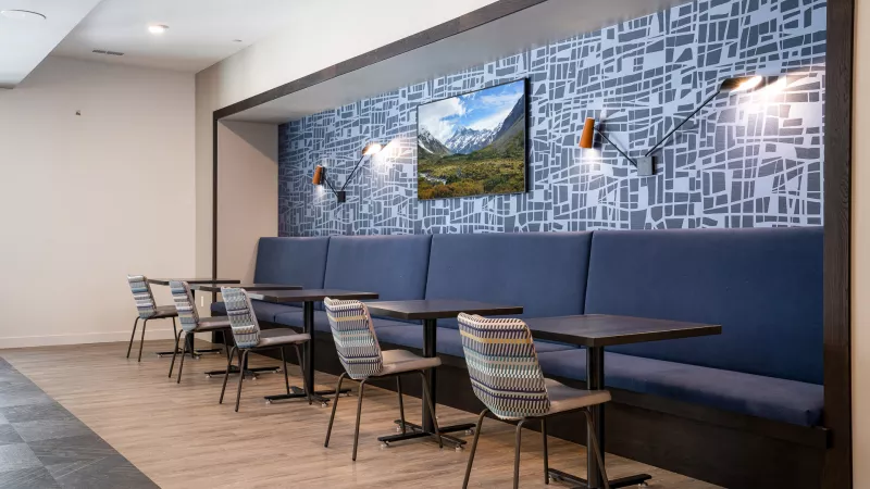Modern dining space with a long blue bench, dark tables, patterned chairs, and a mountain landscape painting on a blue geometric wall.