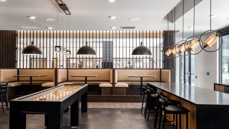 Modern recreational room with shuffleboard, dark counter with stools, and light brown booths under stylish pendant lights.