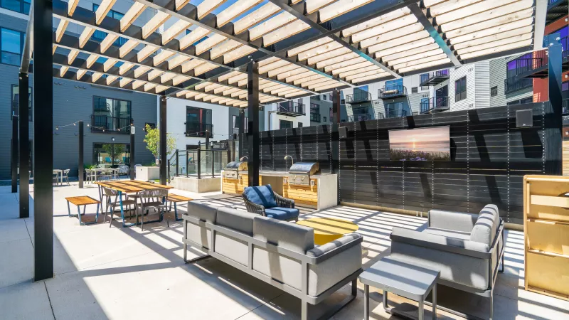 Sunny modern apartment patio with a pergola, grey outdoor sofas, dining area, kitchen, and a large TV on the wall.