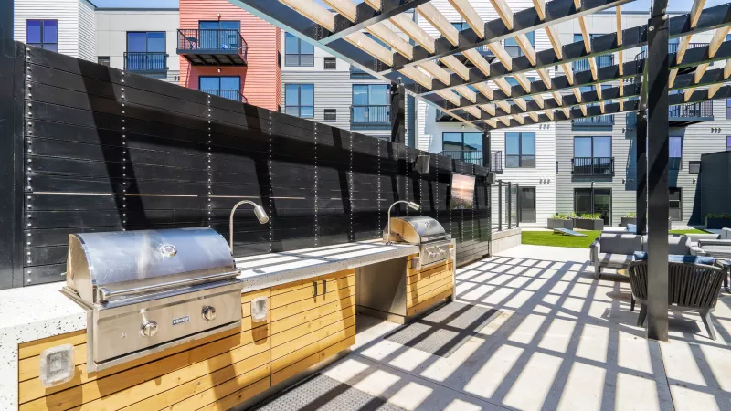 Photograph: Modern outdoor BBQ area with stainless steel grills, a sink, and wooden cabinets, shaded by a slatted pergola.
