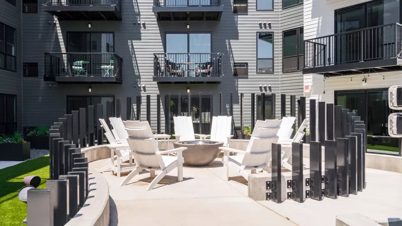 Modern outdoor patio with white lounge chairs, fire pit, and dark curved fence, set against an apartment building.