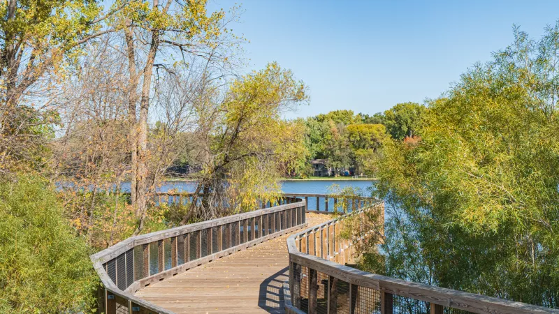 Wooden boardwalk curving over a lake, surrounded by trees with green and autumn foliage under a blue sky.