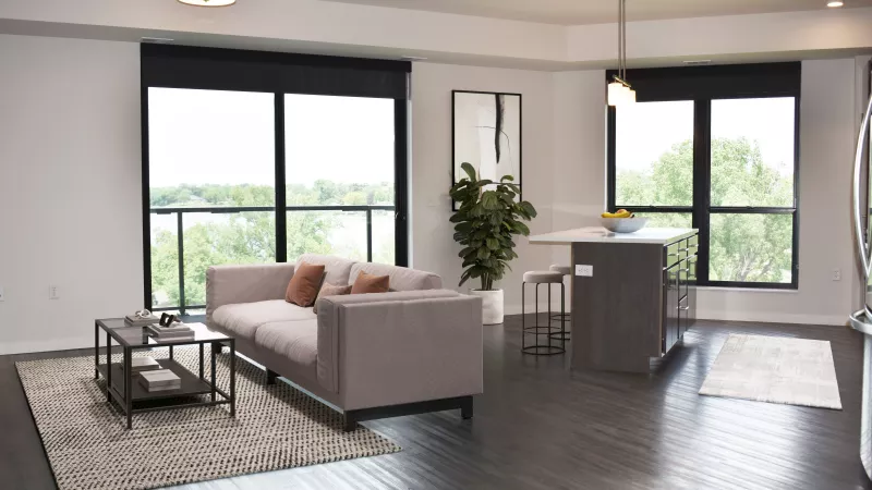 Modern apartment living room with light gray sofa, dark wood floors, large windows, and a kitchen island.