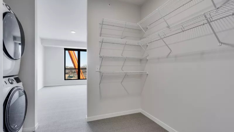Stacked washer and dryer in a laundry closet, beside an empty walk-in closet with wire shelving. Grey carpet, white walls.