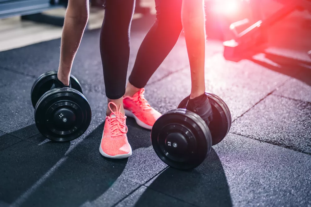 Person in pink sneakers and black leggings bending to lift dumbbells from a dark gym floor, illuminated by red light.