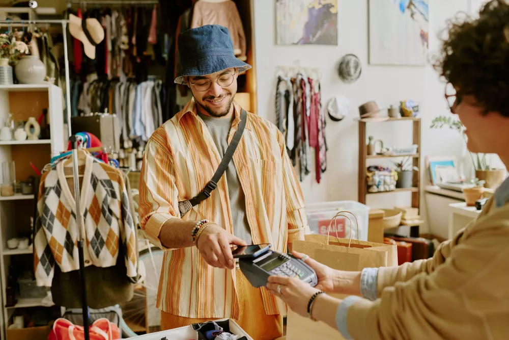 Smiling man in a bucket hat pays with a card at a small boutique. Cashier holds the payment terminal.