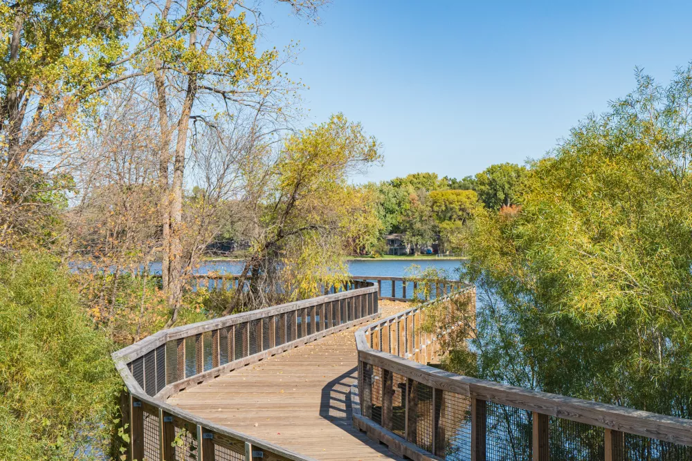 Wooden boardwalk curving over a lake, surrounded by trees with green and autumn foliage under a blue sky.