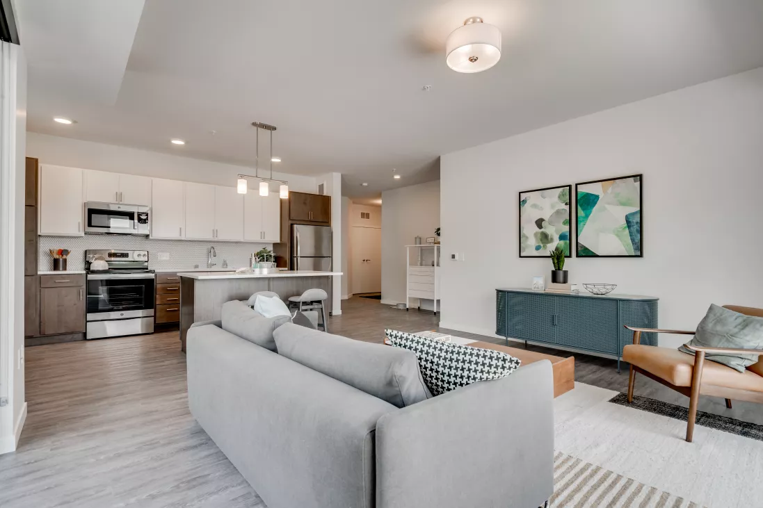 Modern open-plan living room and kitchen. Grey sofa, coffee table, teal console with art, and kitchen island with light cabinets.